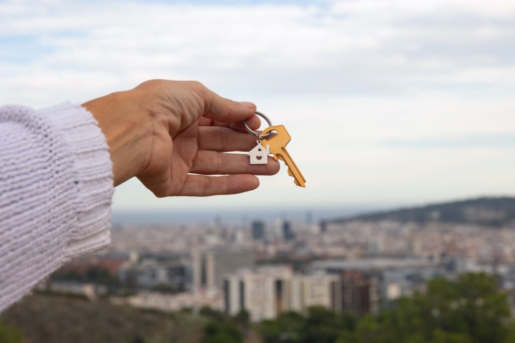 Hand holding a key with a house-shaped keychain, cityscape in the background.