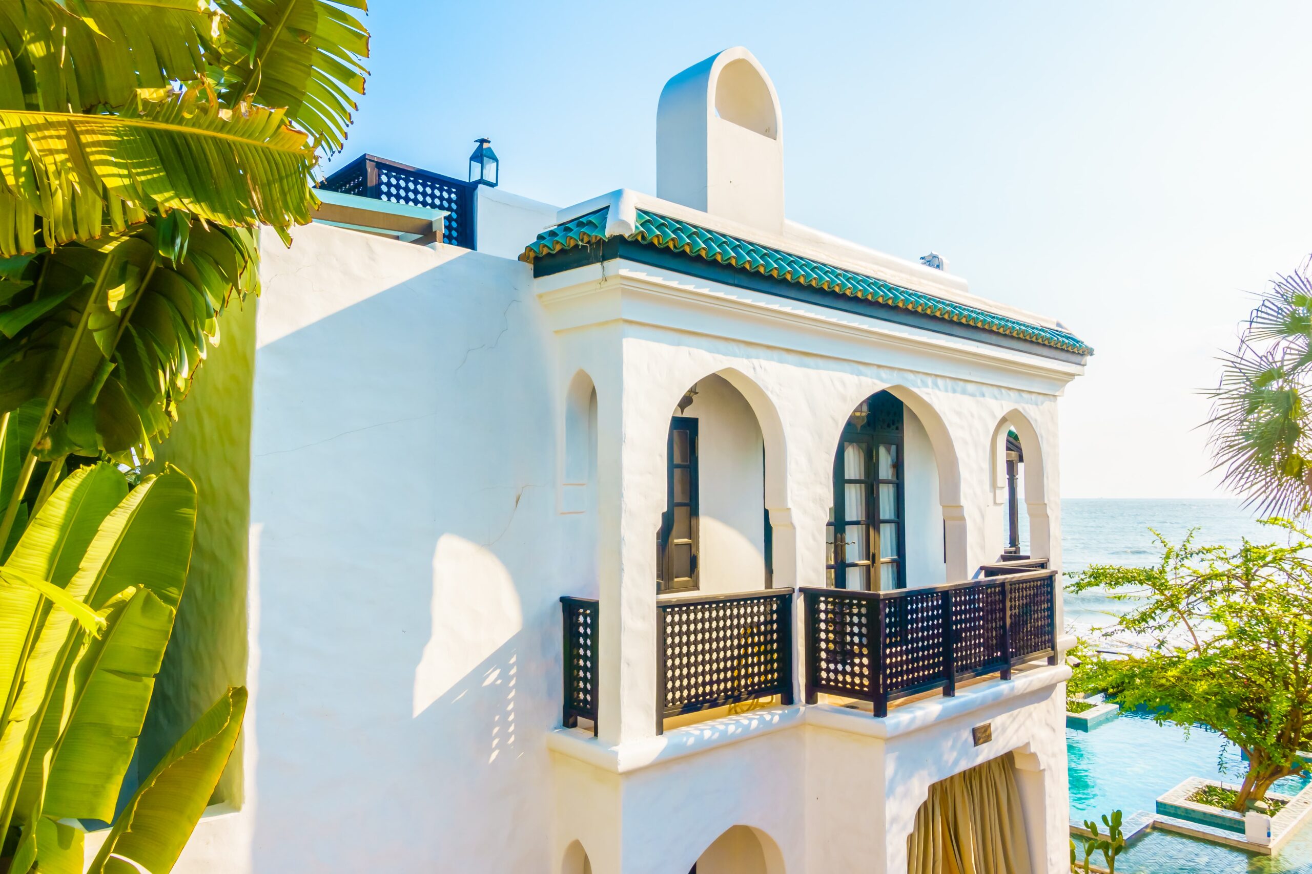 A luxurious white Mediterranean-style villa with arched windows and a balcony, overlooking the sea in Cyprus, surrounded by lush greenery and palm trees.