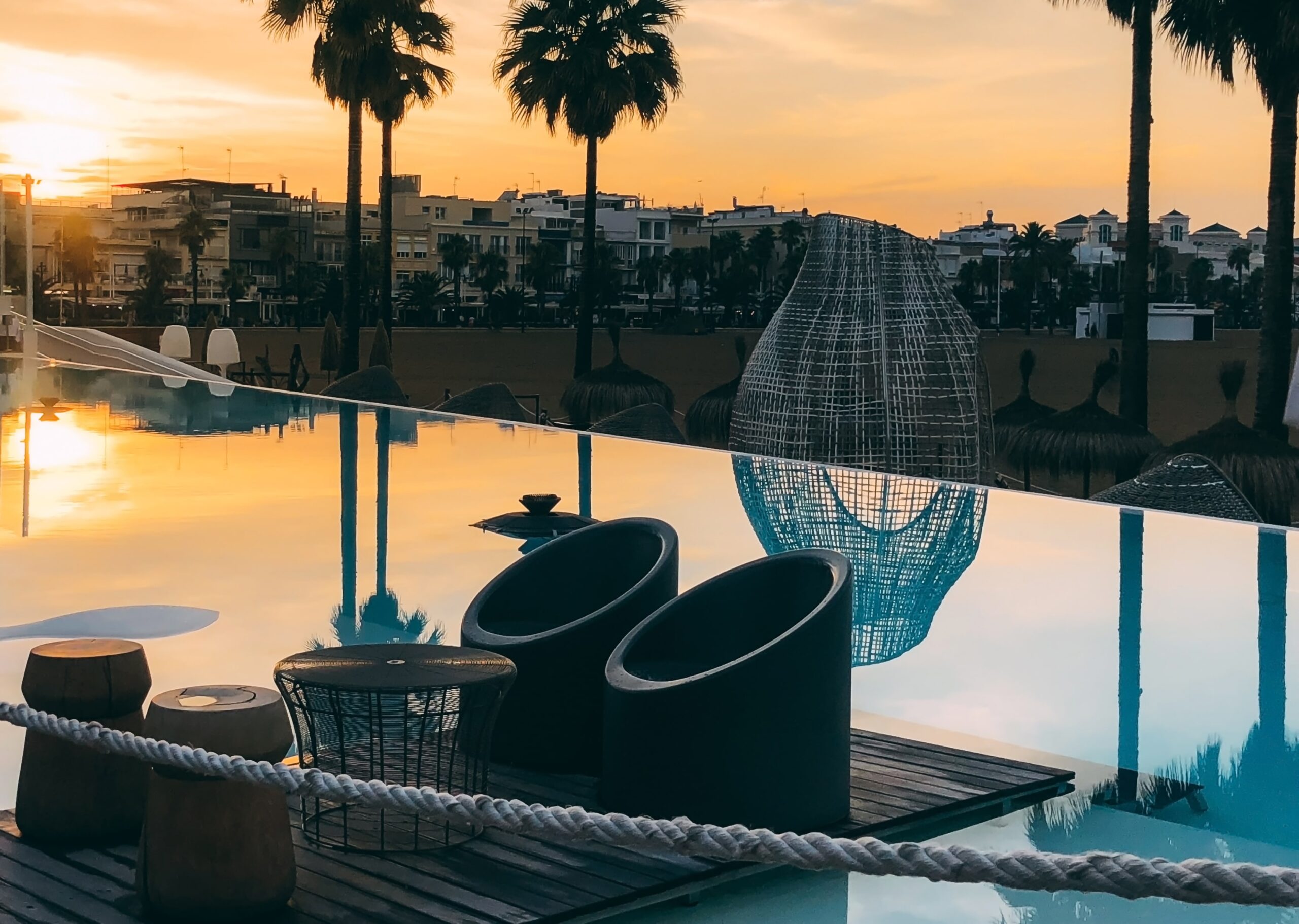 Luxury pool area in Cyprus at sunset, featuring modern outdoor furniture, palm trees, and a calm, reflective water surface.