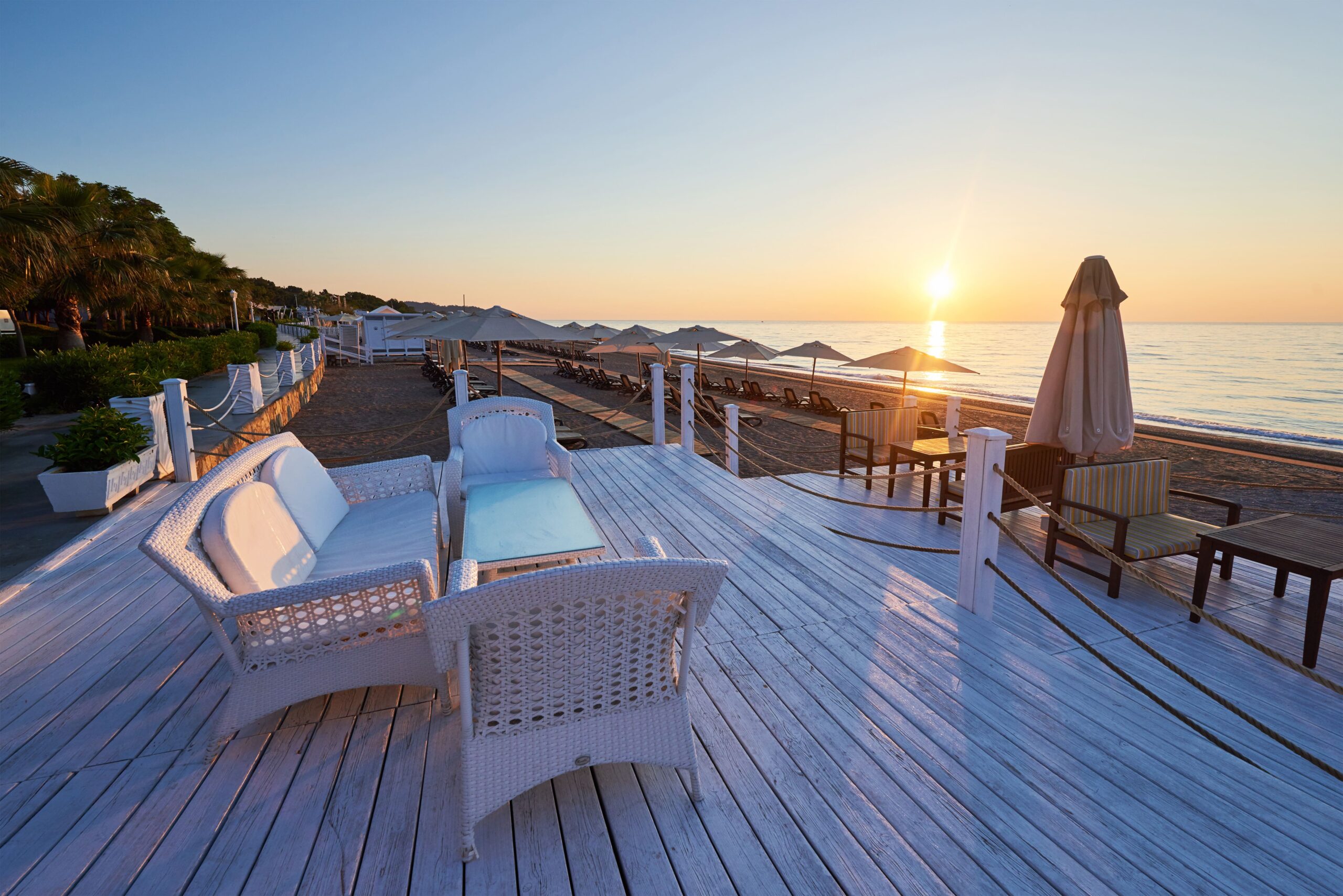 Elegant beachfront seating area in Cyprus with white wicker furniture, overlooking the beach and ocean at sunset.