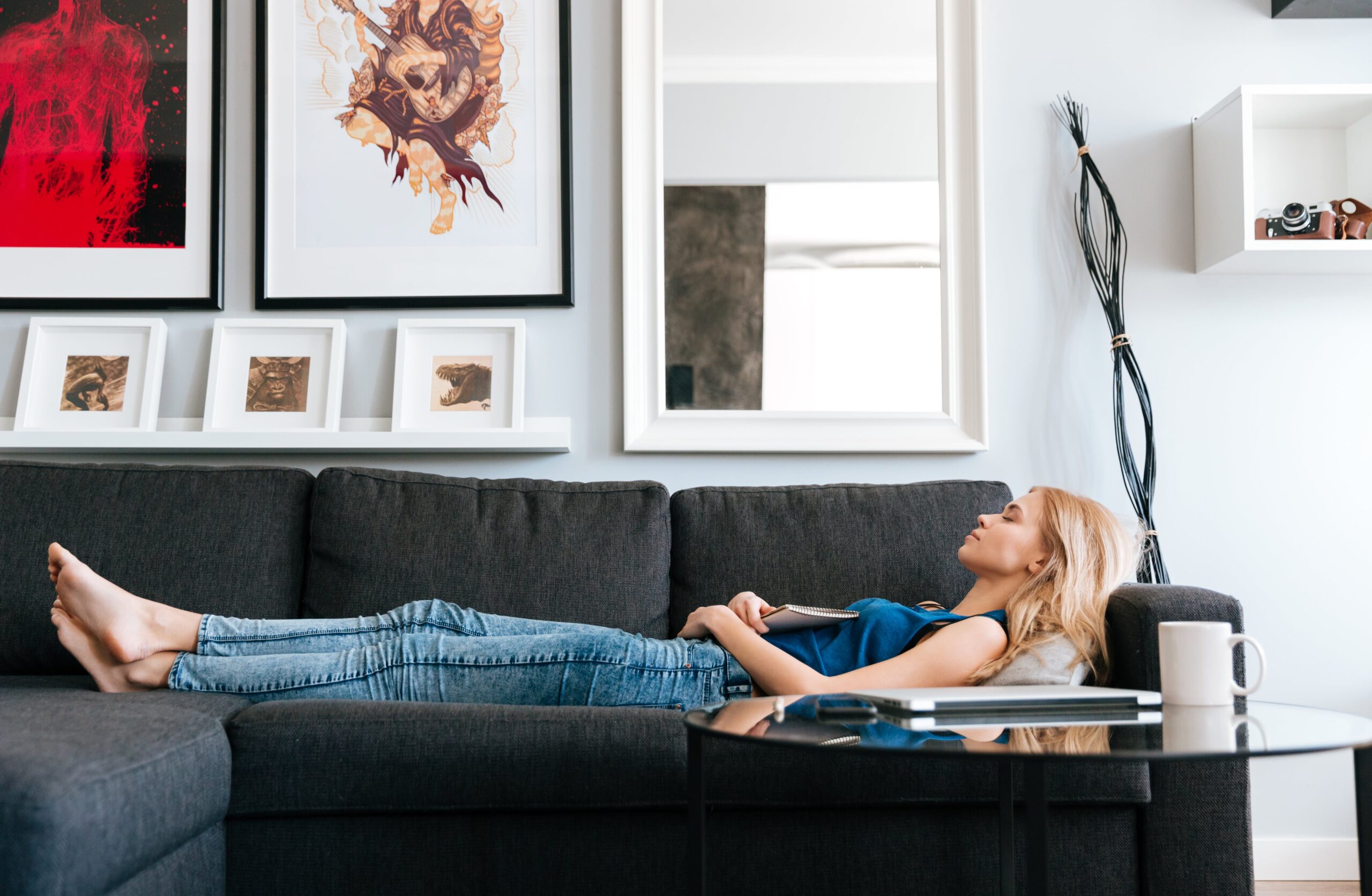 A young woman relaxing on a dark gray sofa in a modern living room, holding a notebook on her lap. The walls are adorned with framed art, and a glass coffee table with a mug and notebook is in the foreground.