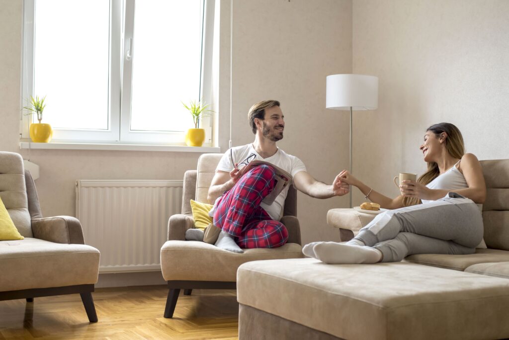 A happy couple sitting in a cozy living room, enjoying a relaxed moment together. The man is wearing red plaid pajama pants, while the woman is holding a coffee mug. The room is decorated with beige furniture, yellow cushions, and potted plants near a bright window.