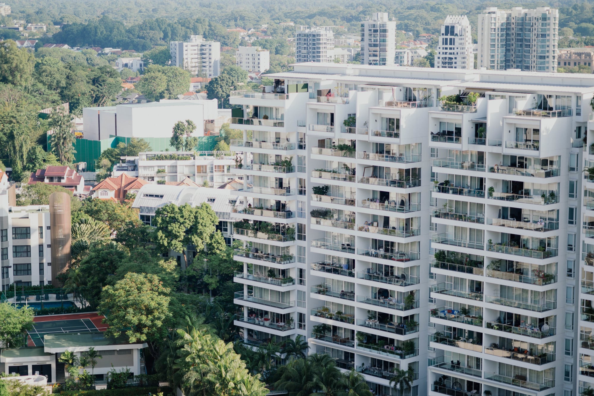 Aerial view of modern white apartment buildings surrounded by lush greenery and nearby residential houses, with a tennis court visible in the foreground.