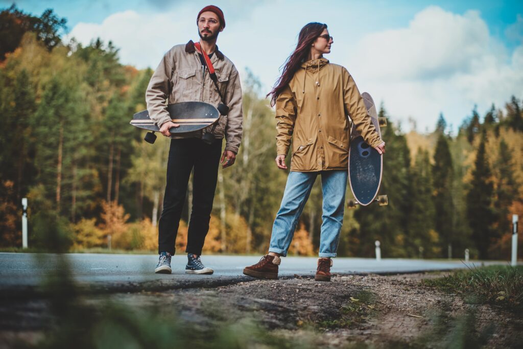 A man and woman holding longboards, standing outdoors on a rural road with a forest in the background.