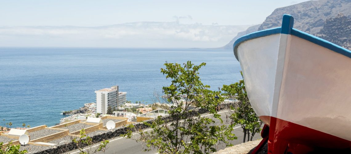 A scenic coastal view in Cyprus featuring a white and red boat on a hillside overlooking the sea, with a modern building and lush greenery in the background.