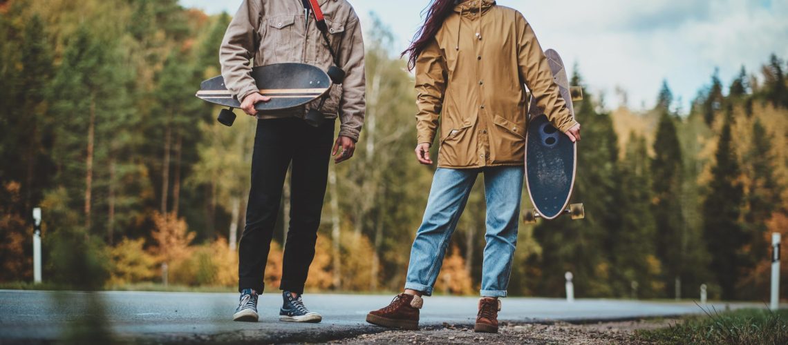 A man and woman holding longboards, standing outdoors on a rural road with a forest in the background.