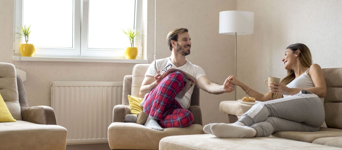 A happy couple sitting in a cozy living room, enjoying a relaxed moment together. The man is wearing red plaid pajama pants, while the woman is holding a coffee mug. The room is decorated with beige furniture, yellow cushions, and potted plants near a bright window.