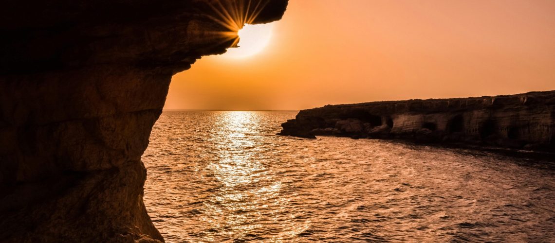 Sunset over Cyprus sea caves with golden light reflecting on the ocean and rocky cliffs.