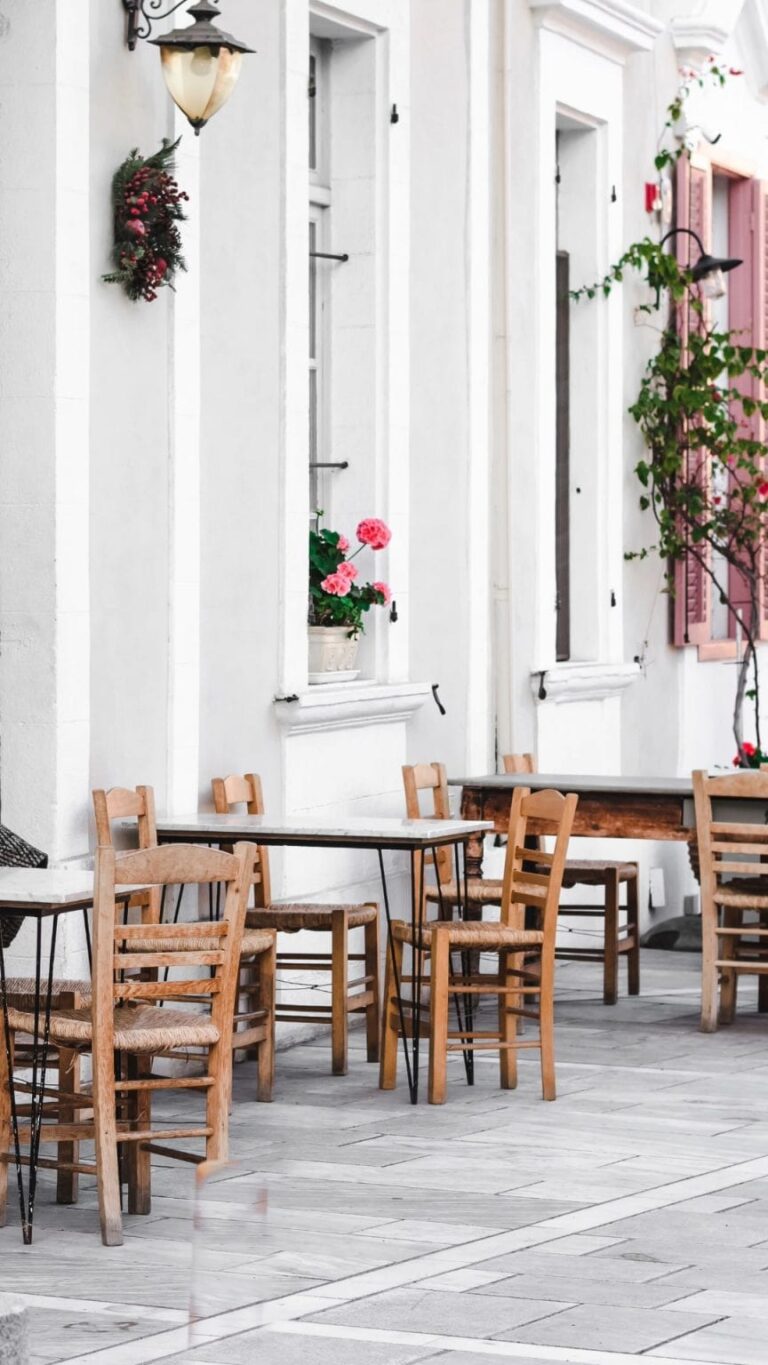 Outdoor café seating area on a bright white Mediterranean-style street in Larnaca, Cyprus, featuring wooden chairs, potted flowers, and traditional architecture.