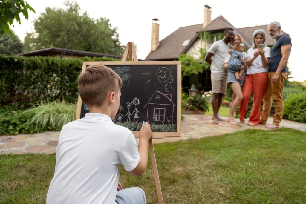 Child drawing a house on a chalkboard in a garden while a family stands together in the background near a home.
