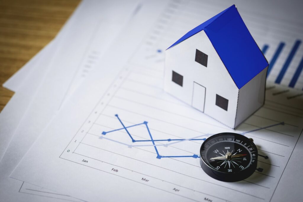 Model house with blue roof placed on financial charts next to a compass, illustrating a house for sale in Larnaca and real estate market trends.